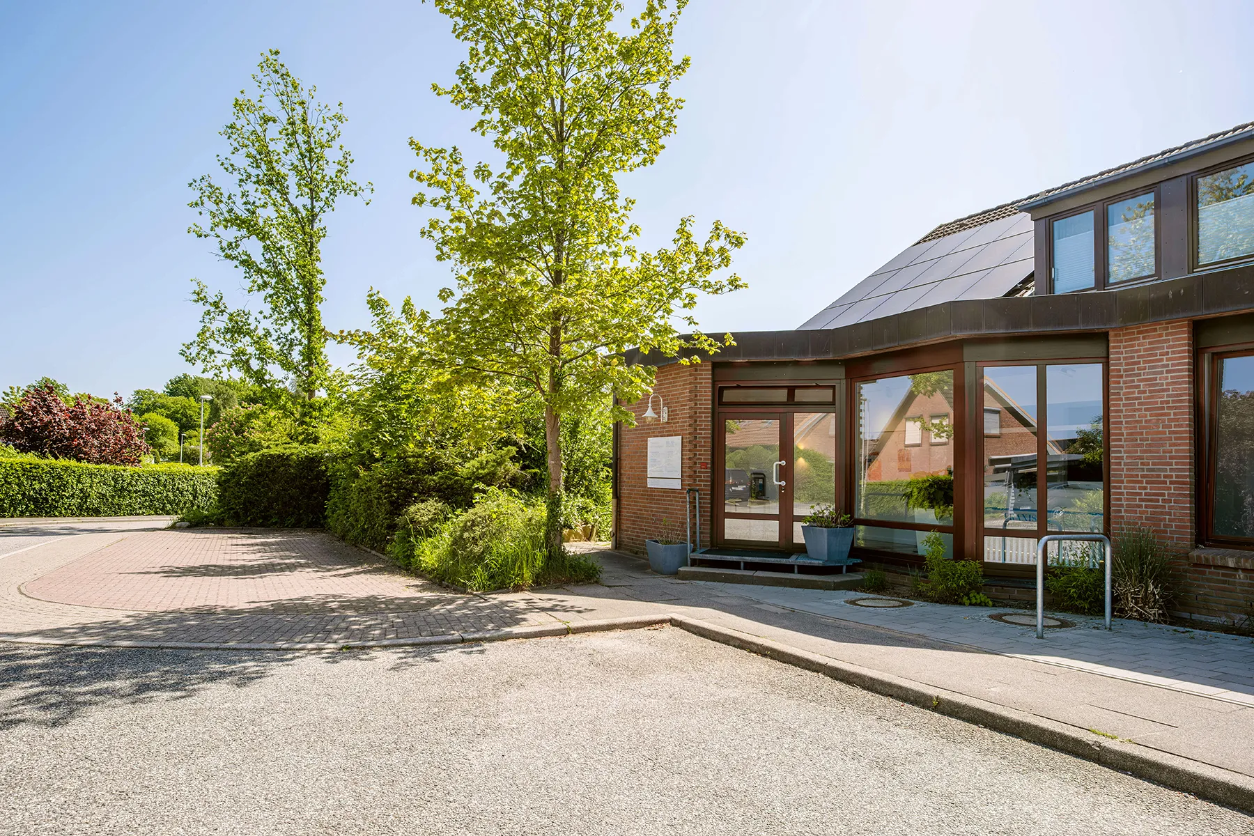 Praxis Barkauer Kontakt Vor Ort Ein modernes Gebäude mit Ziegelfassade, großen Fenstern und Solarpanelen auf dem Dach. Zwei Bäume und Büsche stehen davor. Der Himmel ist sonnig und klar. Im Vordergrund ist ein leerer Parkplatz.
