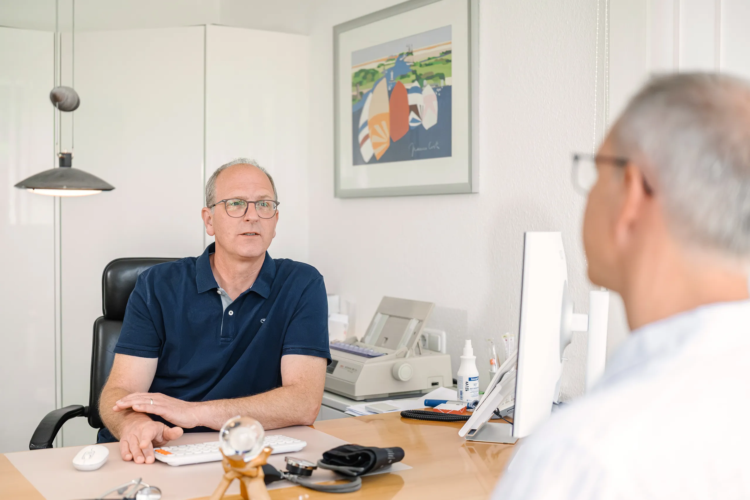 Ein älterer Mann in einem blauen Shirt sitzt an einem Schreibtisch und spricht mit einer anderen Person, die ihm gegenüber sitzt. Der Raum ist hell, mit einem dekorativen Bild an der Wand und einem Computer auf dem Tisch.