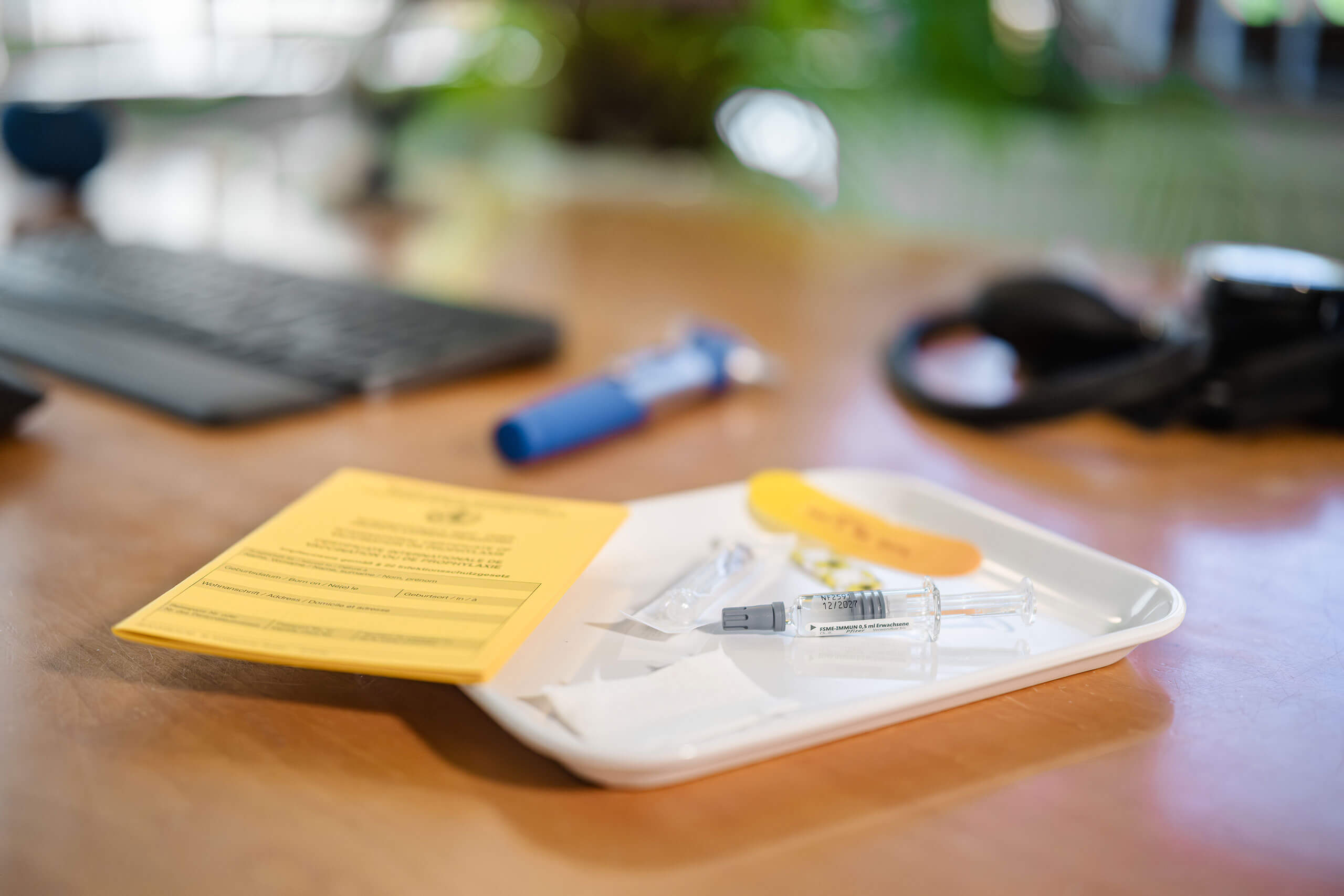 Praxis Barkauer Strasse Kiel Arbeitsmedizin 5 Ein Impfausweis liegt auf einem Tablett neben einer Spritze und einer durchsichtigen Ampulle auf einem Schreibtisch. Im Hintergrund sind unscharf eine Tastatur und Kopfhörer zu erkennen.