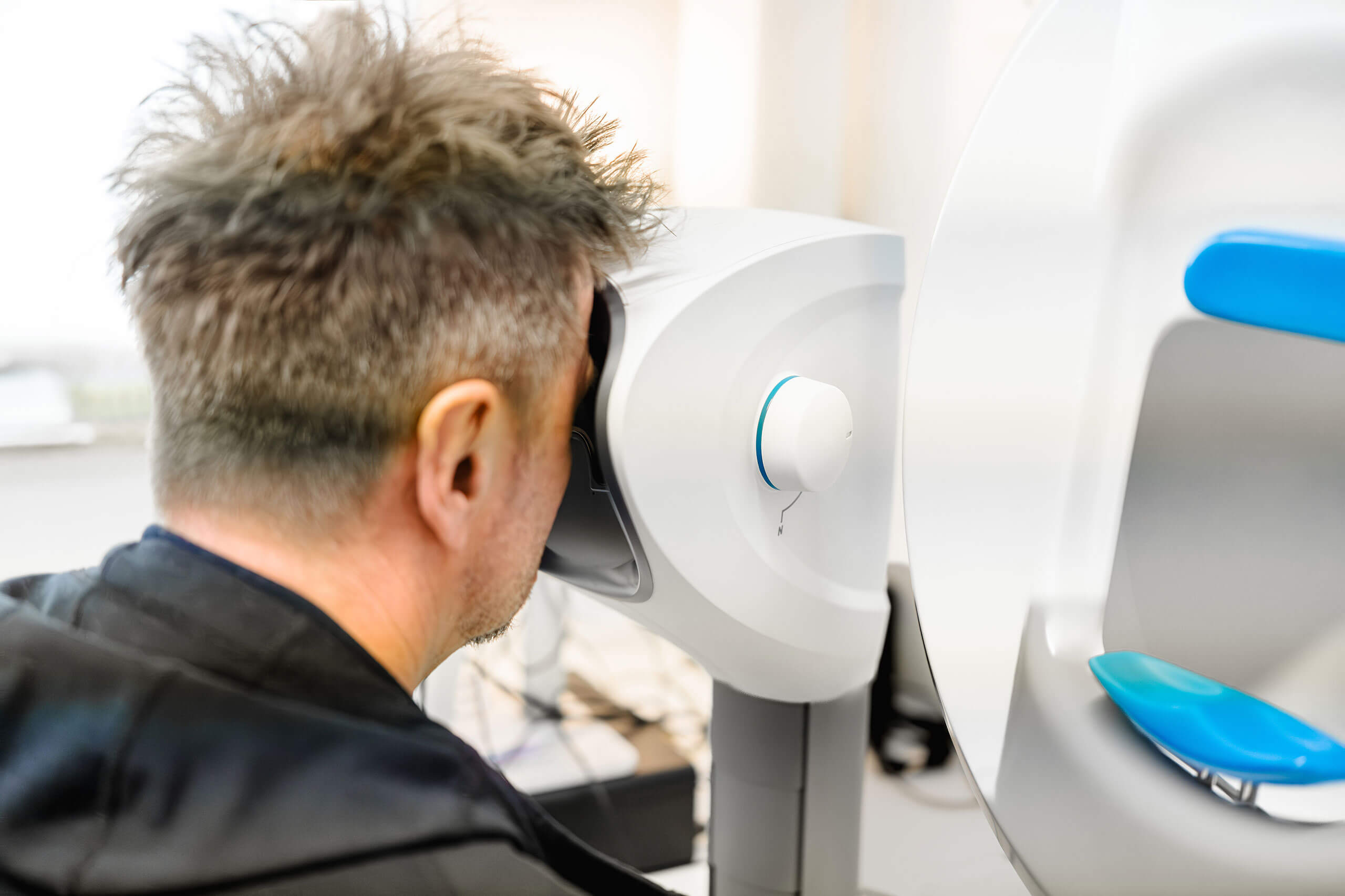 Praxis Barkauer Strasse Kiel Arbeitsmedizin 6 Eine Person sitzt vor einem ophthalmologischen Gerät und schaut in eine Öffnung für eine Augenuntersuchung. Das Gerät ist weiß mit einem blauen Akzent und steht in einem hellen Raum.
