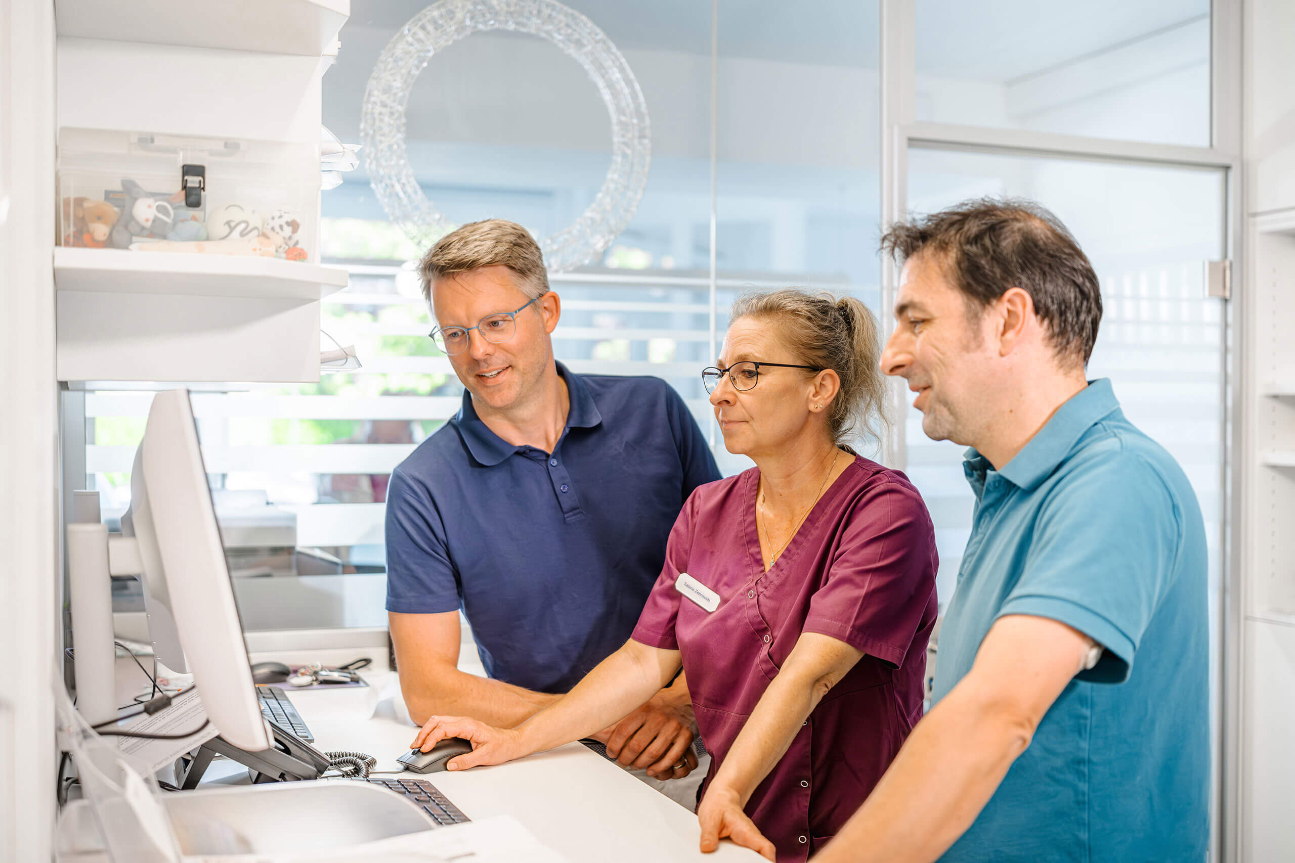 Drei Personen stehen an einem Schreibtisch und schauen auf einen Computerbildschirm. Sie tragen Freizeit- oder Berufskleidung. Im Hintergrund ist ein Büro mit Glaswänden zu sehen.