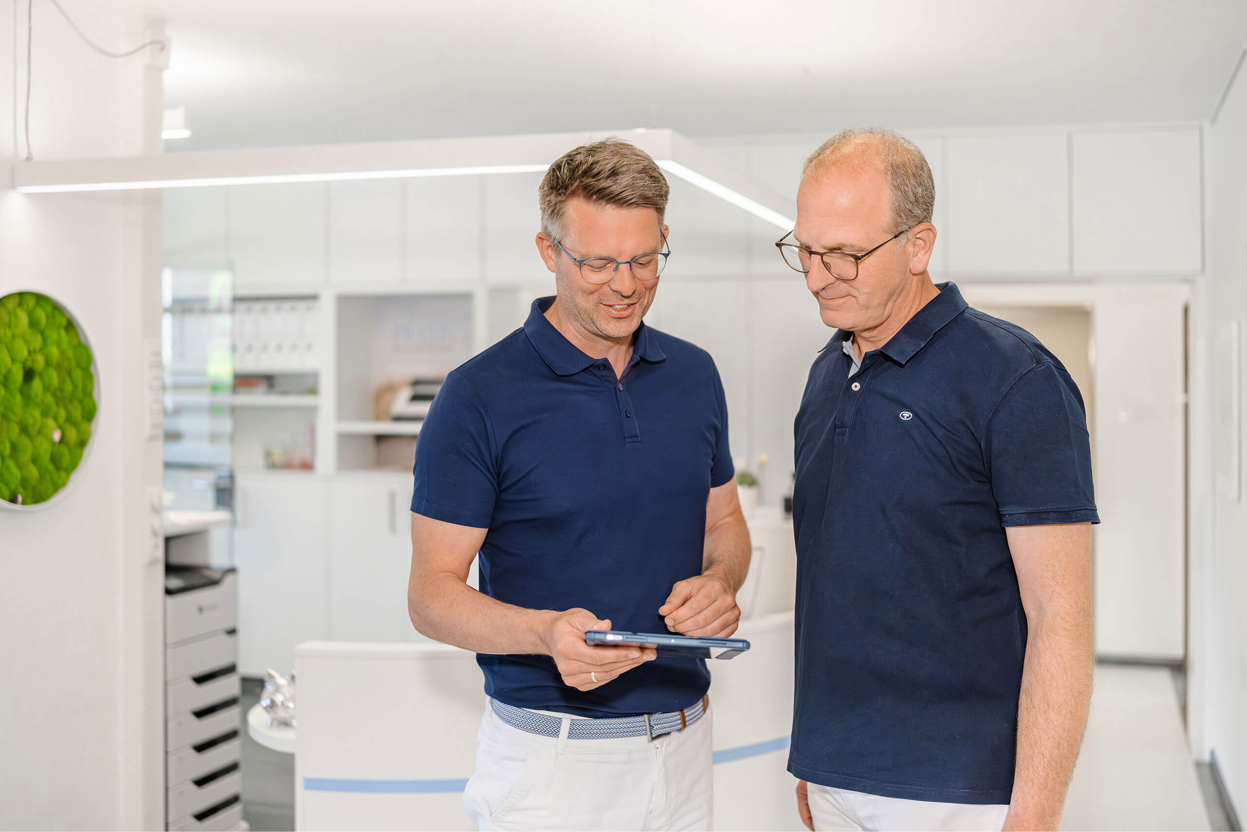 Zwei Männer stehen in einem Büro und betrachten gemeinsam ein Tablet. Beide tragen blaue Polohemden. Im Hintergrund sind weiße Schränke und ein runder, grüner Pflanzenrahmen an der Wand zu sehen.