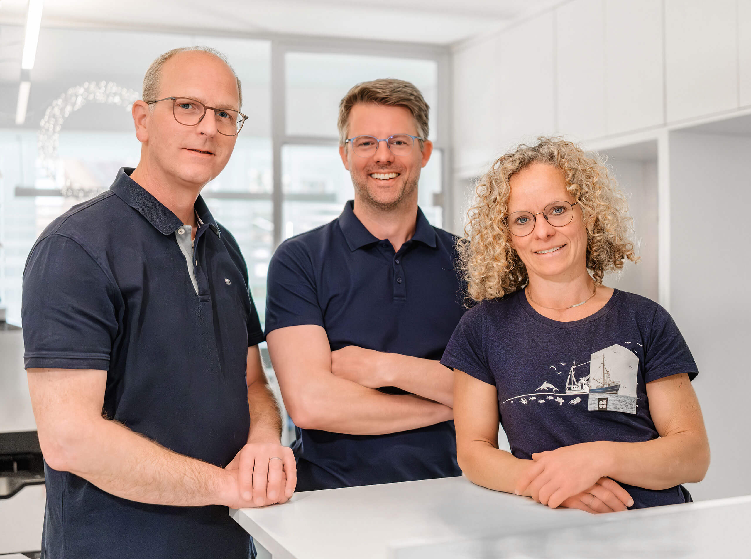 Drei Personen stehen in einem hellen Büro. Zwei Männer und eine Frau lächeln in die Kamera. Alle tragen Brillen und dunkelblaue Oberteile. Sie stehen hinter einem weißen Tisch. Im Hintergrund sind Fenster und weiße Schränke.