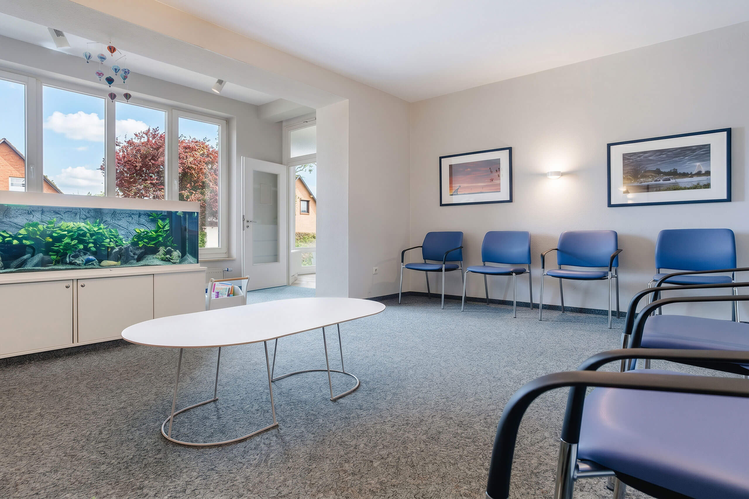 Ein Wartezimmer mit vier blauen Stühlen an der Wand, einem weißen Tisch in der Mitte, einem großen Aquarium links und zwei gerahmten Bildern an der Wand. Tageslicht strömt durch ein Fenster hinein.