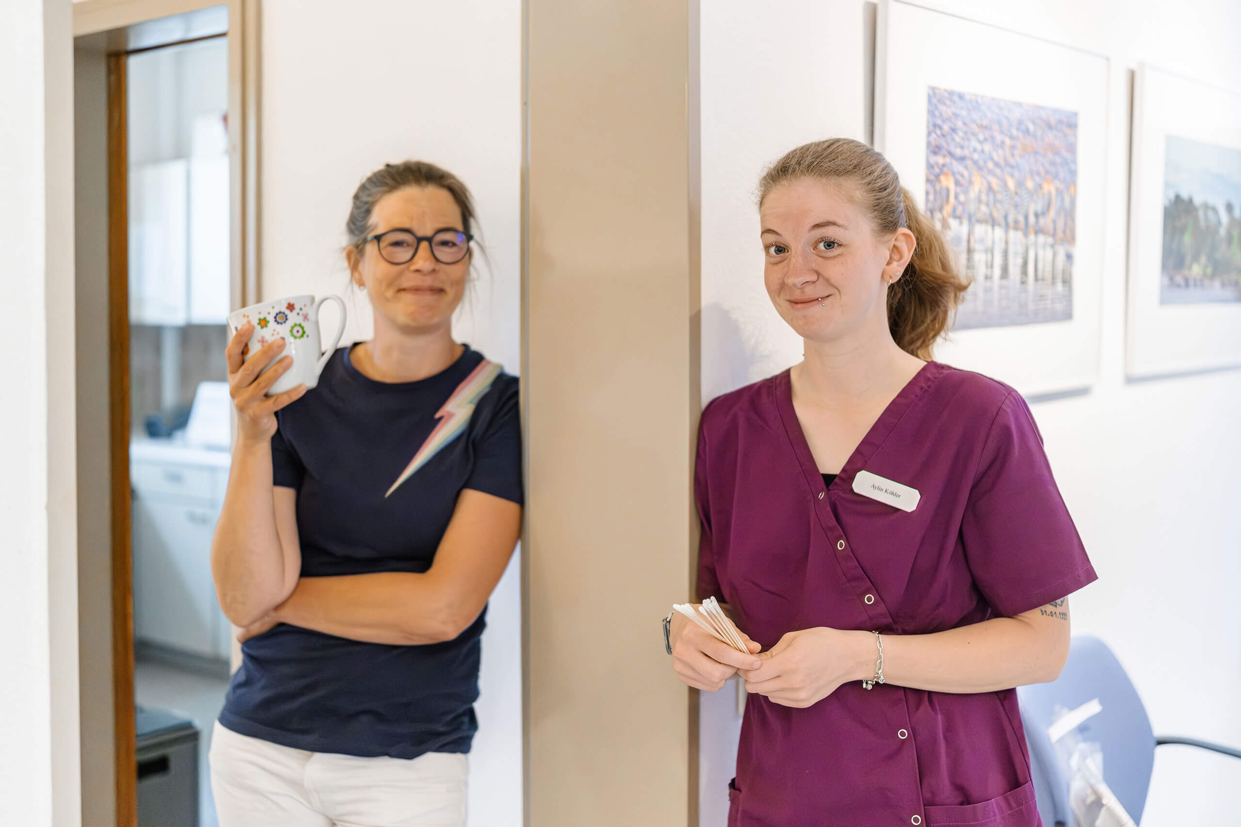 Zwei Personen stehen in einem Innenraum. Die linke Person hält eine Tasse und trägt eine Brille, die rechte Person trägt ein lilafarbenes Oberteil. Beide lächeln und stehen vor einer Wand mit gerahmten Bildern.