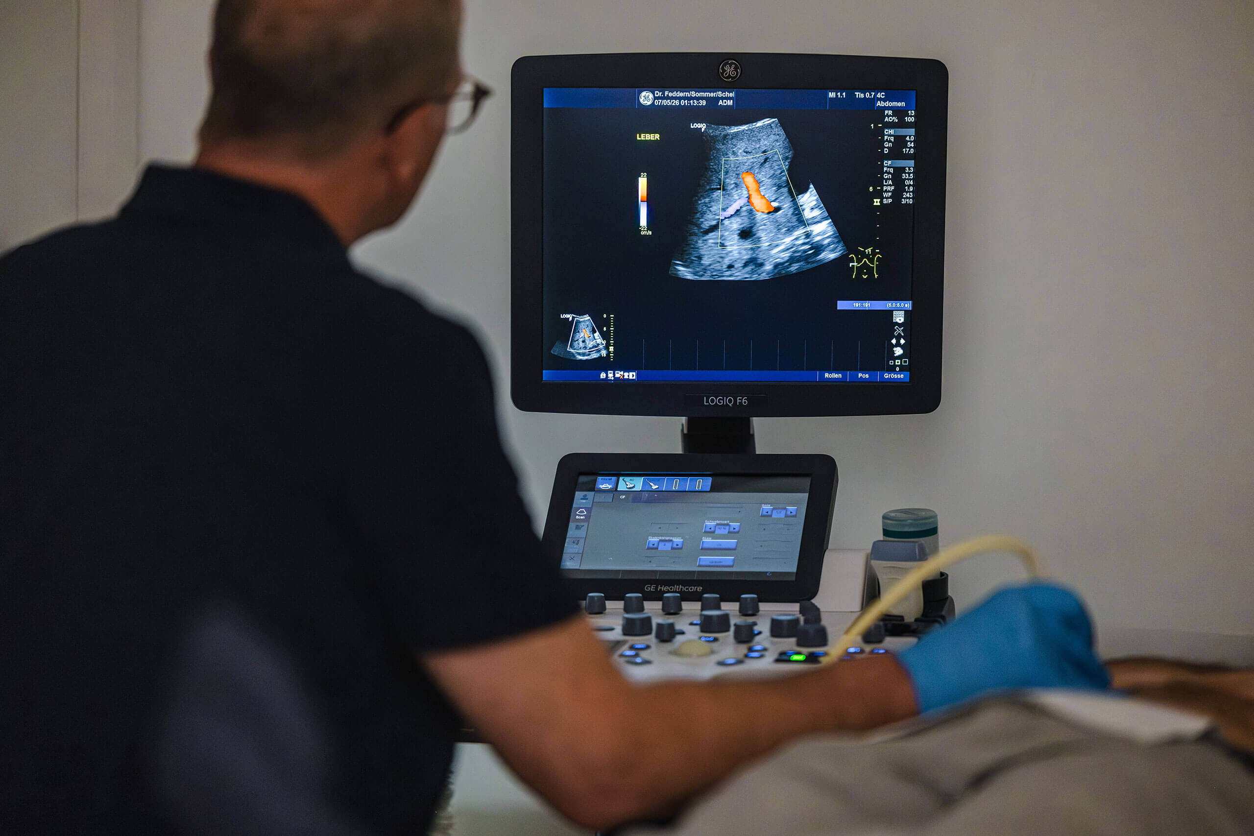 Praxis Barkauer Strasse Kiel Hausaerztliche Versorgung 2 Ein Arzt führt einen Ultraschall durch. Der Bildschirm zeigt ein Schwarz-Weiß-Bild eines Organs mit einem farbigen Bereich. Der Arzt trägt Handschuhe und hält das Ultraschallgerät. Im Hintergrund sind Bedienelemente sichtbar.